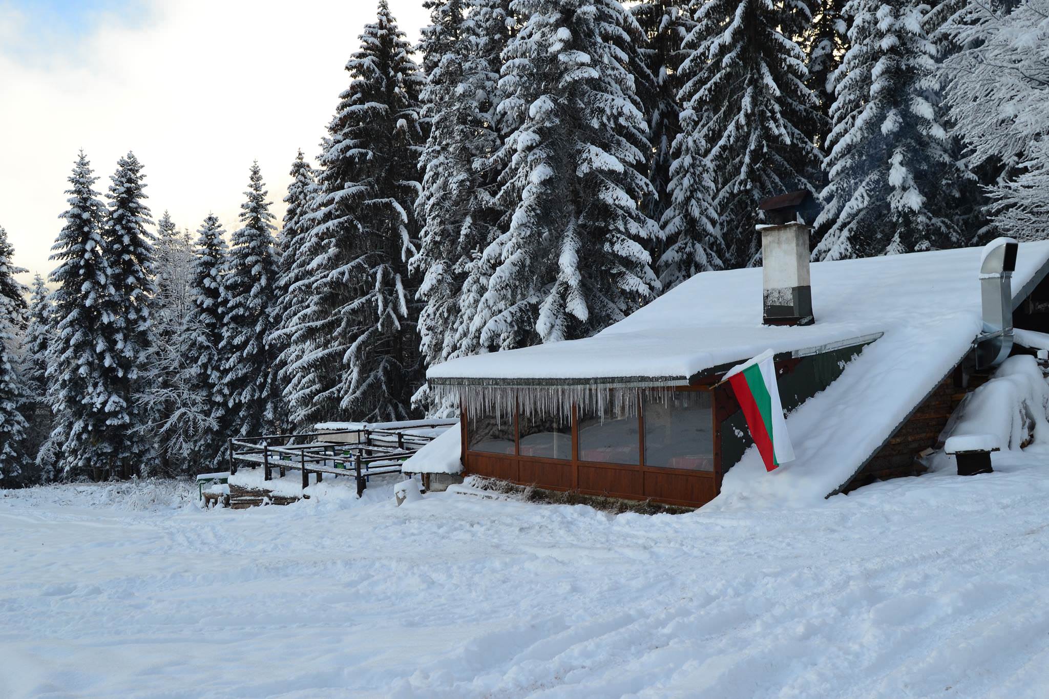 Photo of the teahouse outside. Small wooden house with smoking chimney nestled in the snow. To the left is a trellis with benches and tables. On the right side of the house is the Bulgarian flag.